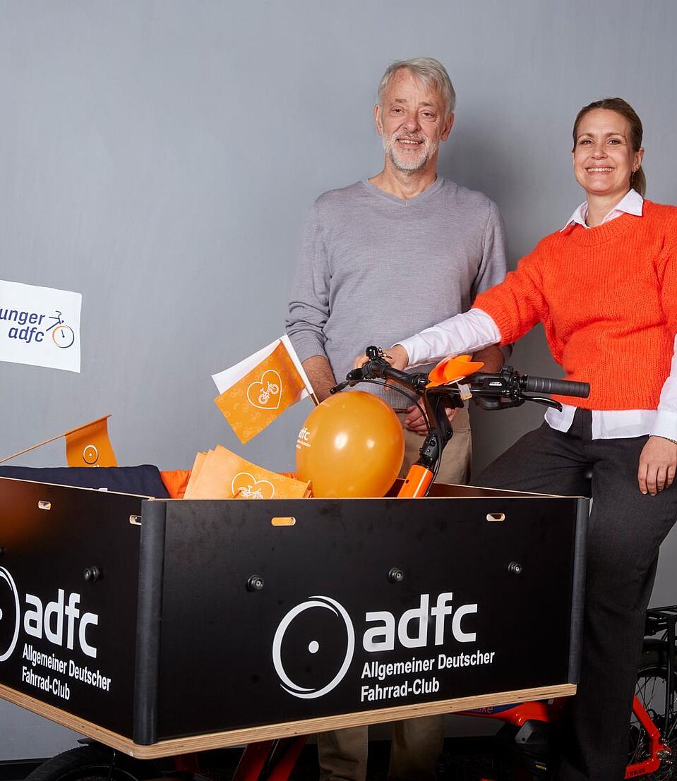 Holczer und Masurat Frank Masurat steht hinter einem Lastenrad, auf dem Sarah Holczer sitzt. Im Hintergrund eine blau-weiße ADFC-Fotowand.